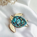 A handcrafted sea turtle brooch with a gold outline, cream-colored flippers, and a blue and green mosaic-patterned shell is displayed on white satin fabric with dried grasses nearby.