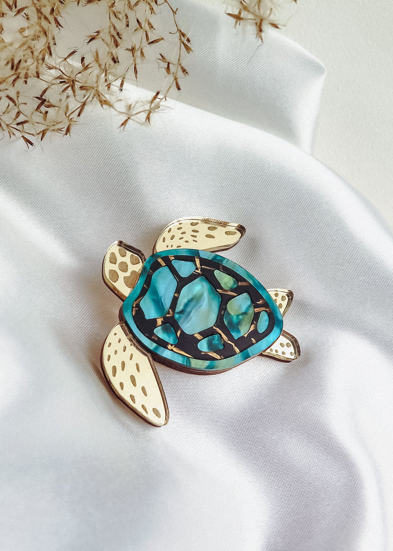 A handcrafted sea turtle brooch with a gold outline, cream-colored flippers, and a blue and green mosaic-patterned shell is displayed on white satin fabric with dried grasses nearby.