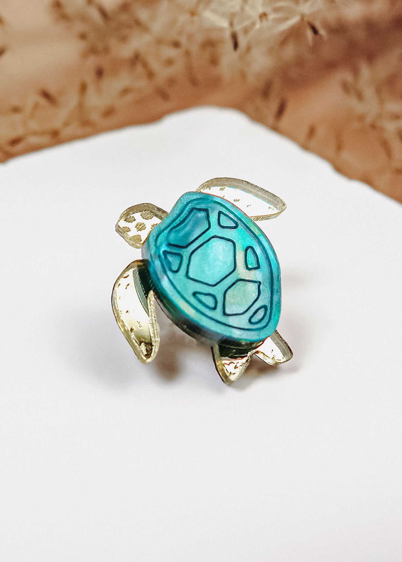 A gold and turquoise sea turtle-shaped ring, handcrafted with mirror-acrylic glass, rests on a white surface against a blurred brown background.
