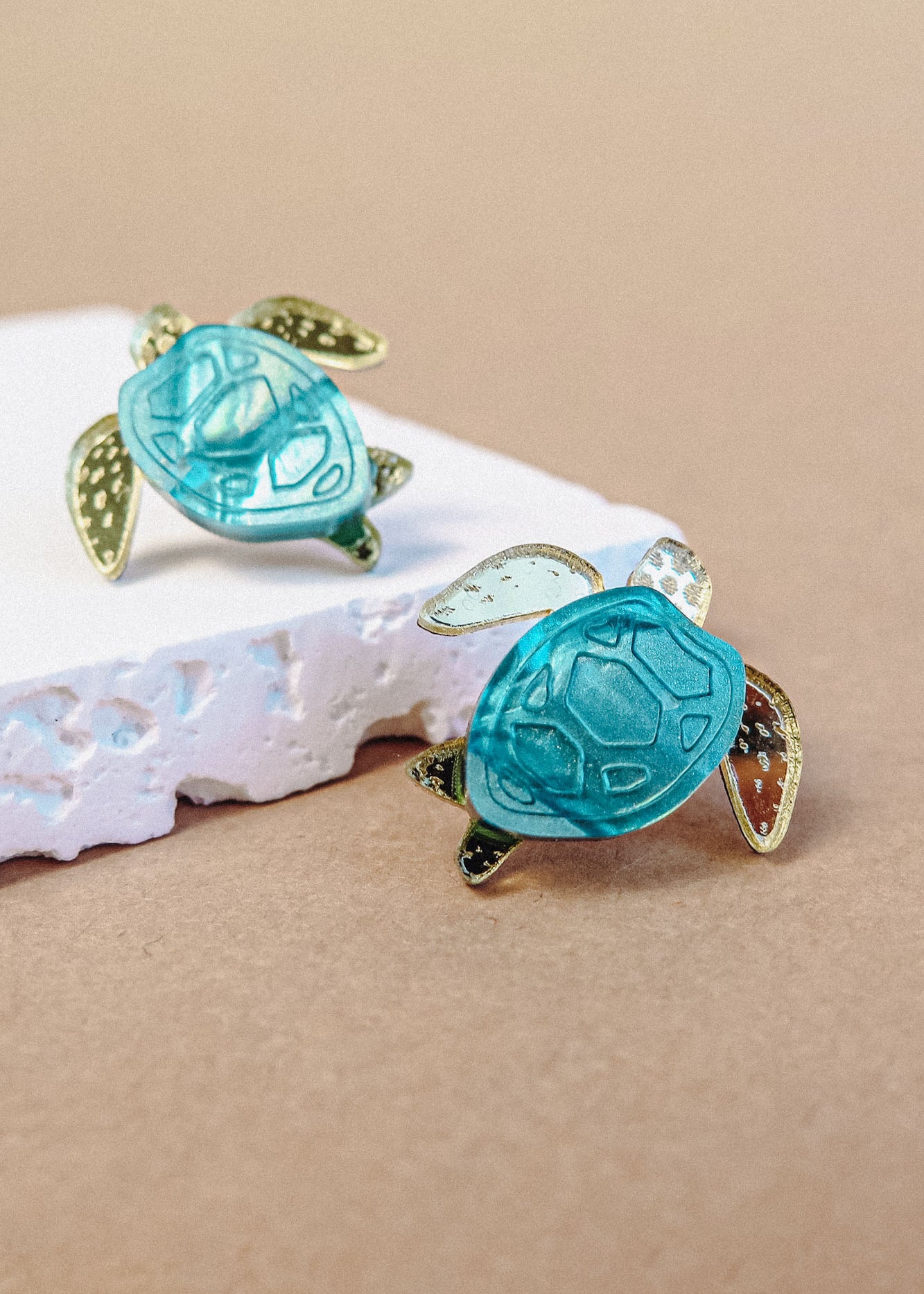 Two gold and turquoise Sea Turtle Studs rest on a piece of textured white material against a beige background, bringing an ocean-inspired, summery vibe to your accessories.