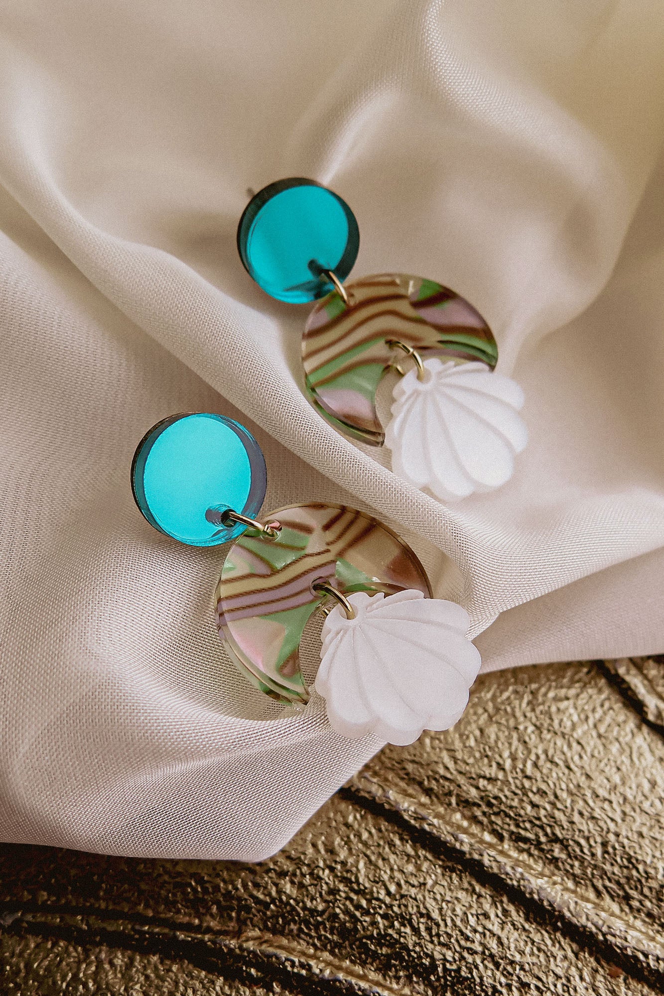A pair of handmade Seashell Earrings featuring round turquoise studs, marbled circular pieces in green, pink, and clear tones, and lightweight white seashell-shaped charms, displayed on draped white fabric.