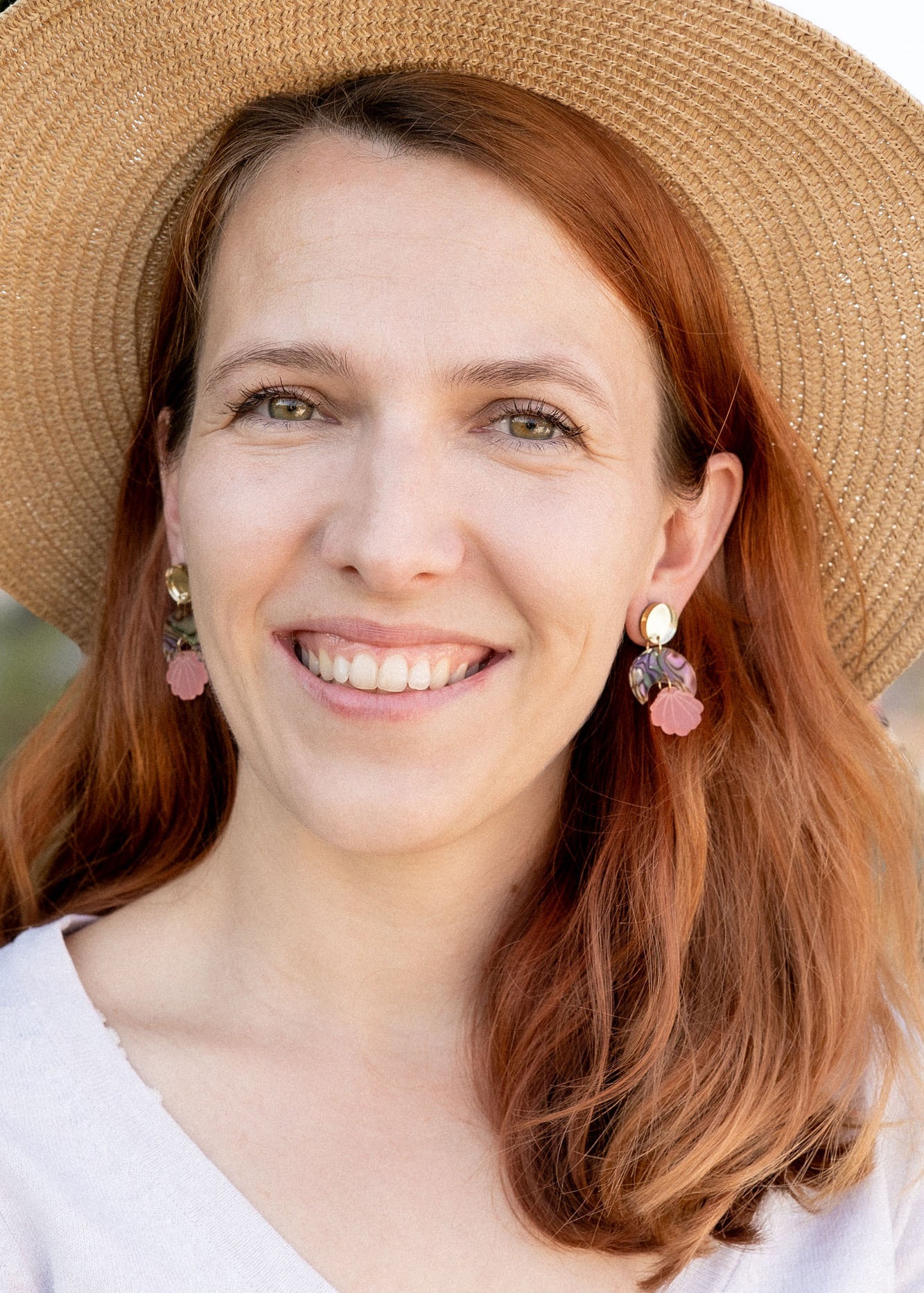 A woman with long auburn hair wearing a wide-brimmed straw hat, lightweight seashell earrings, and a light V-neck top smiles outdoors with greenery in the background.