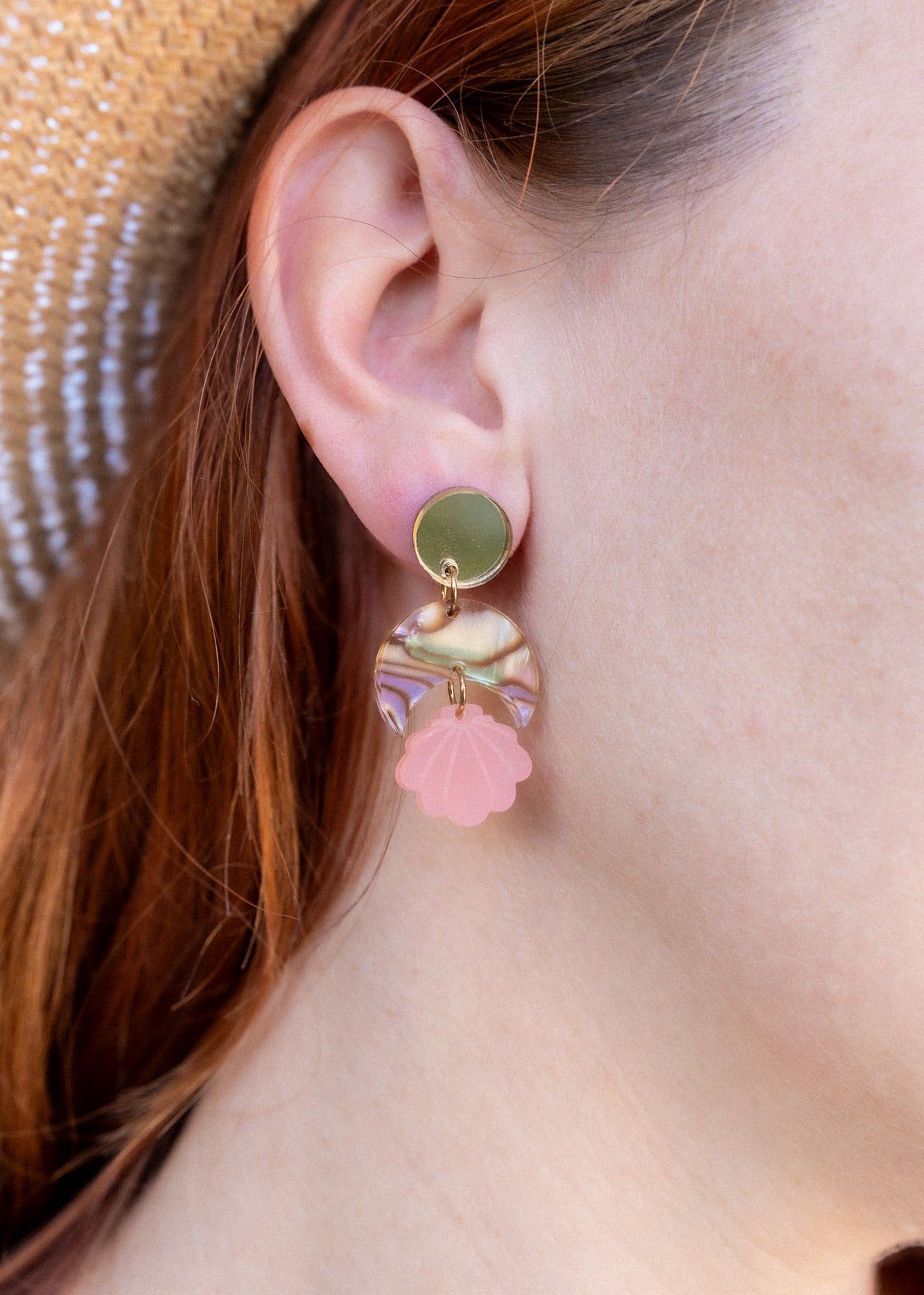 A close-up of a womans ear wearing a handmade gold stud earring with a bow and a dangling pink seashell-shaped charm. She has red hair, is wearing a straw hat, and the lightweight design adds delicate charm.