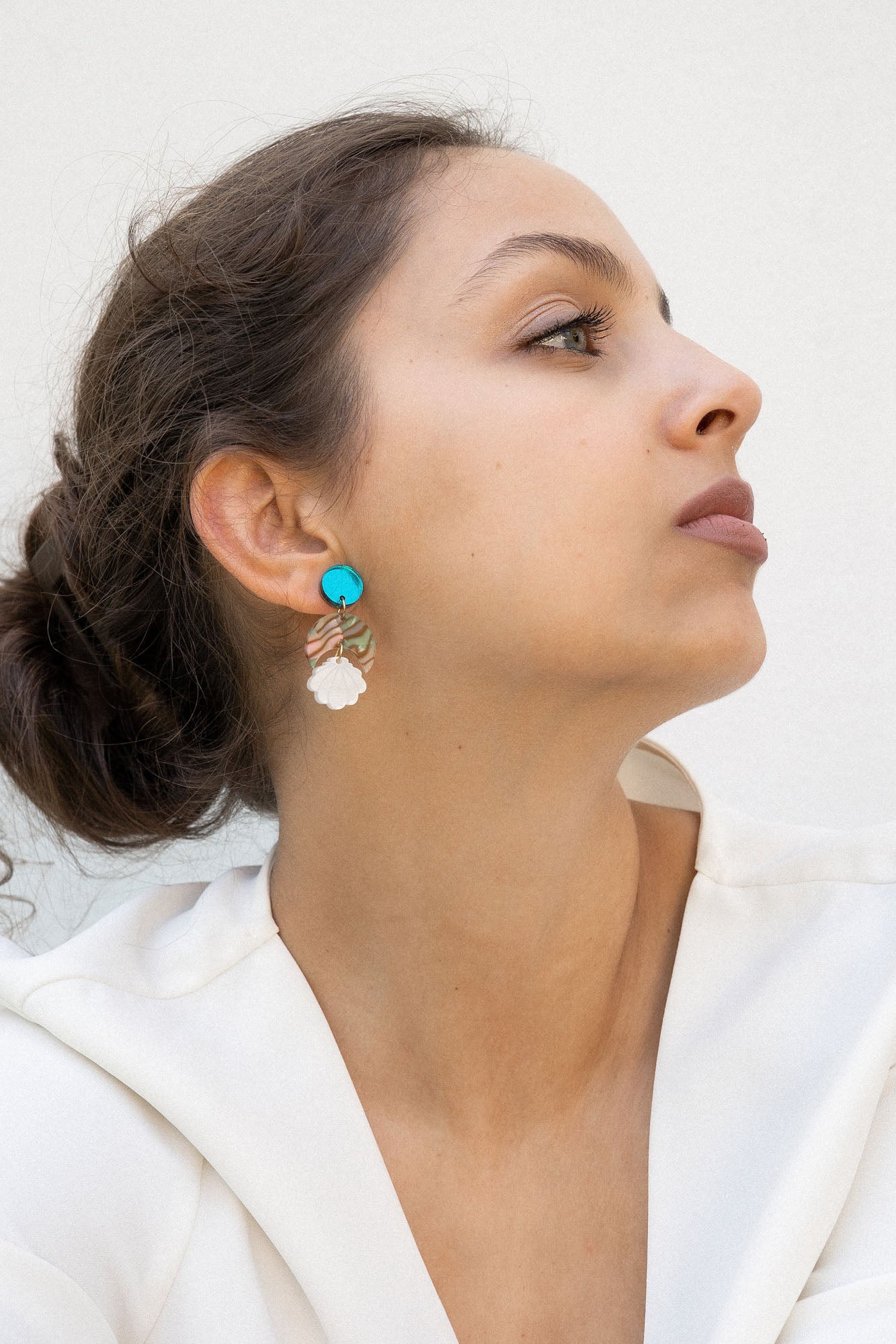 A woman with brown hair in a loose bun wears a white blouse and lightweight statement earrings with turquoise and gold accents. She gazes to the side against a plain, light background.