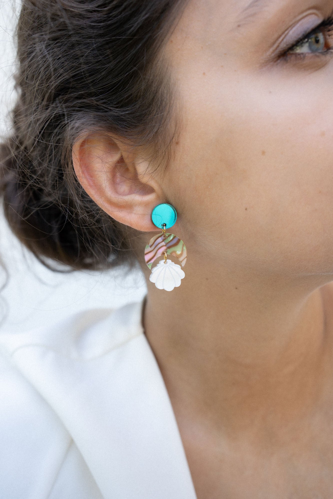Close-up of a woman’s ear wearing a handmade, colorful earring with a turquoise stud, wavy striped section, and white floral-shaped dangle. Her hair is pulled back and she is wearing a white top. Lightweight for all-day comfort.