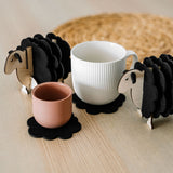 A white mug and a pink cup rest on felt coasters, accompanied by charming wooden sheep coaster holders on a light wooden table. Perfect as home decor accents, with a woven placemat in the background.