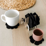 A white mug and a pink cup sit on black felt coasters next to a wooden sheep-shaped coaster holder on a light wooden table with a woven placemat in the background, perfect for charming home decor.