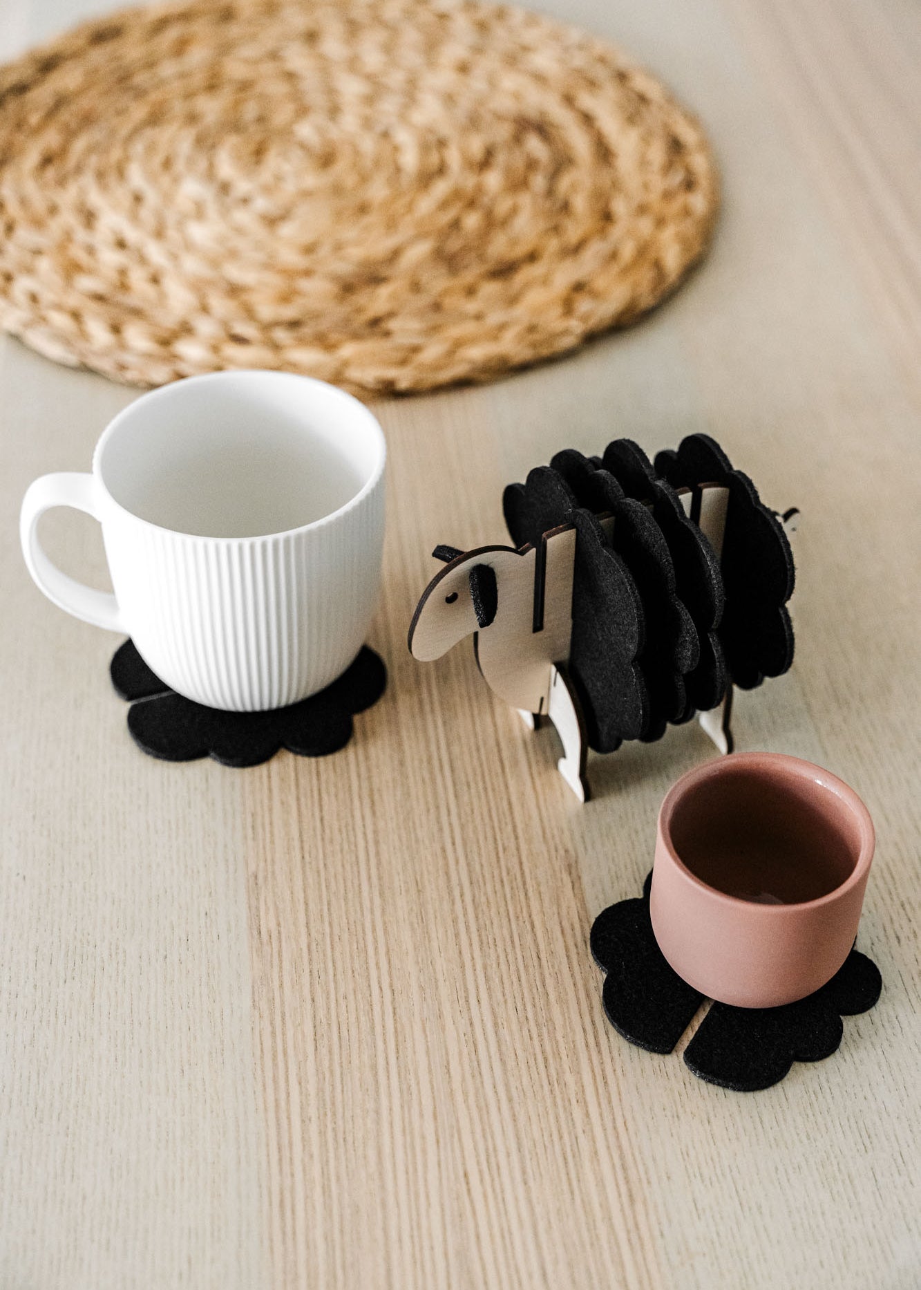A white mug and a pink cup sit on black felt coasters next to a wooden sheep-shaped coaster holder on a light wooden table with a woven placemat in the background, perfect for charming home decor.