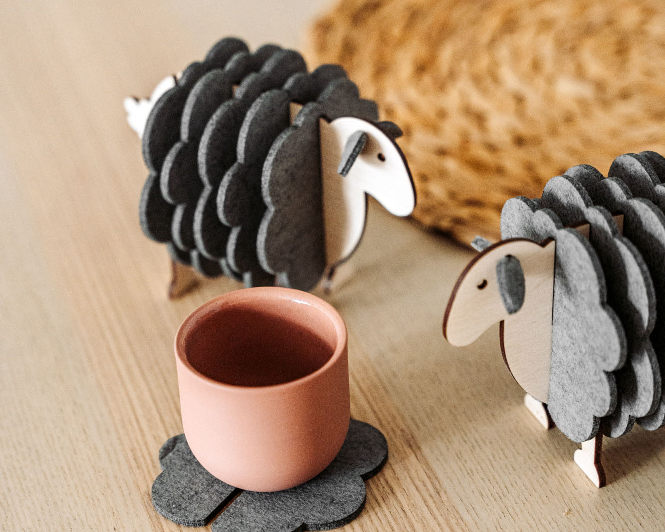A pink cup rests on a gray, flower-shaped felt coaster beside two wooden sheep figurines—charming home decor—placed on a light wooden surface, with a woven mat partially visible in the background.