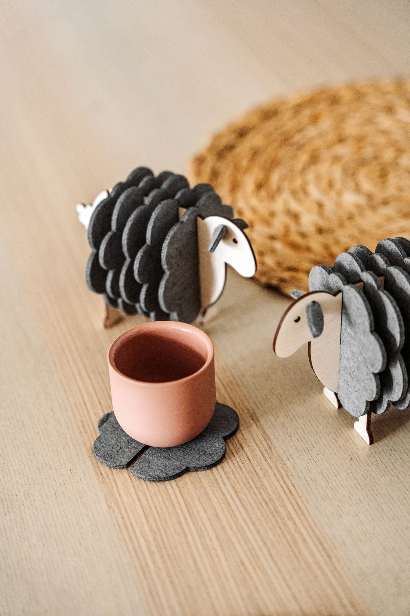 A pink cup rests on a gray, flower-shaped felt coaster beside two wooden sheep figurines—charming home decor—placed on a light wooden surface, with a woven mat partially visible in the background.