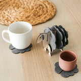 A white mug and a pink cup sit on dark felt coasters next to a decorative wooden sheep-shaped coaster holder, bringing charming home decor touches to a light wooden table topped with a round woven placemat.