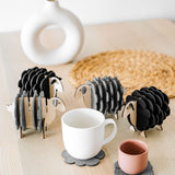 A white table holds a white vase, a woven placemat, and several sheep-shaped felt coasters. A white mug and a pink cup rest on two of the coasters, bringing playful charm to this cozy home decor setting.