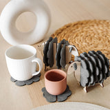 Two wooden sheep-shaped coaster holders with dark gray felt coasters add a touch of charming home decor to a light wooden table, paired with a white mug, pink cup, woven placemat, and modern white vase with dried stems.