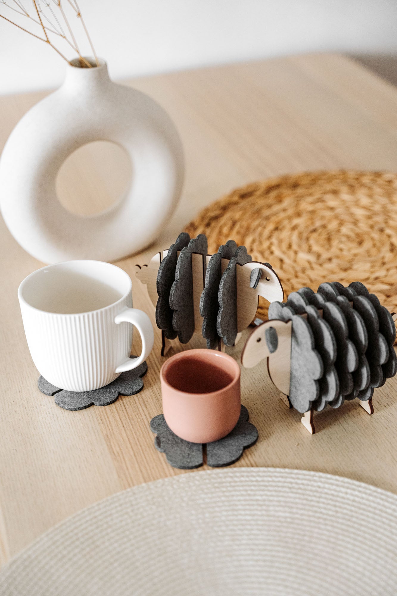 Two wooden sheep-shaped coaster holders with dark gray felt coasters add a touch of charming home decor to a light wooden table, paired with a white mug, pink cup, woven placemat, and modern white vase with dried stems.