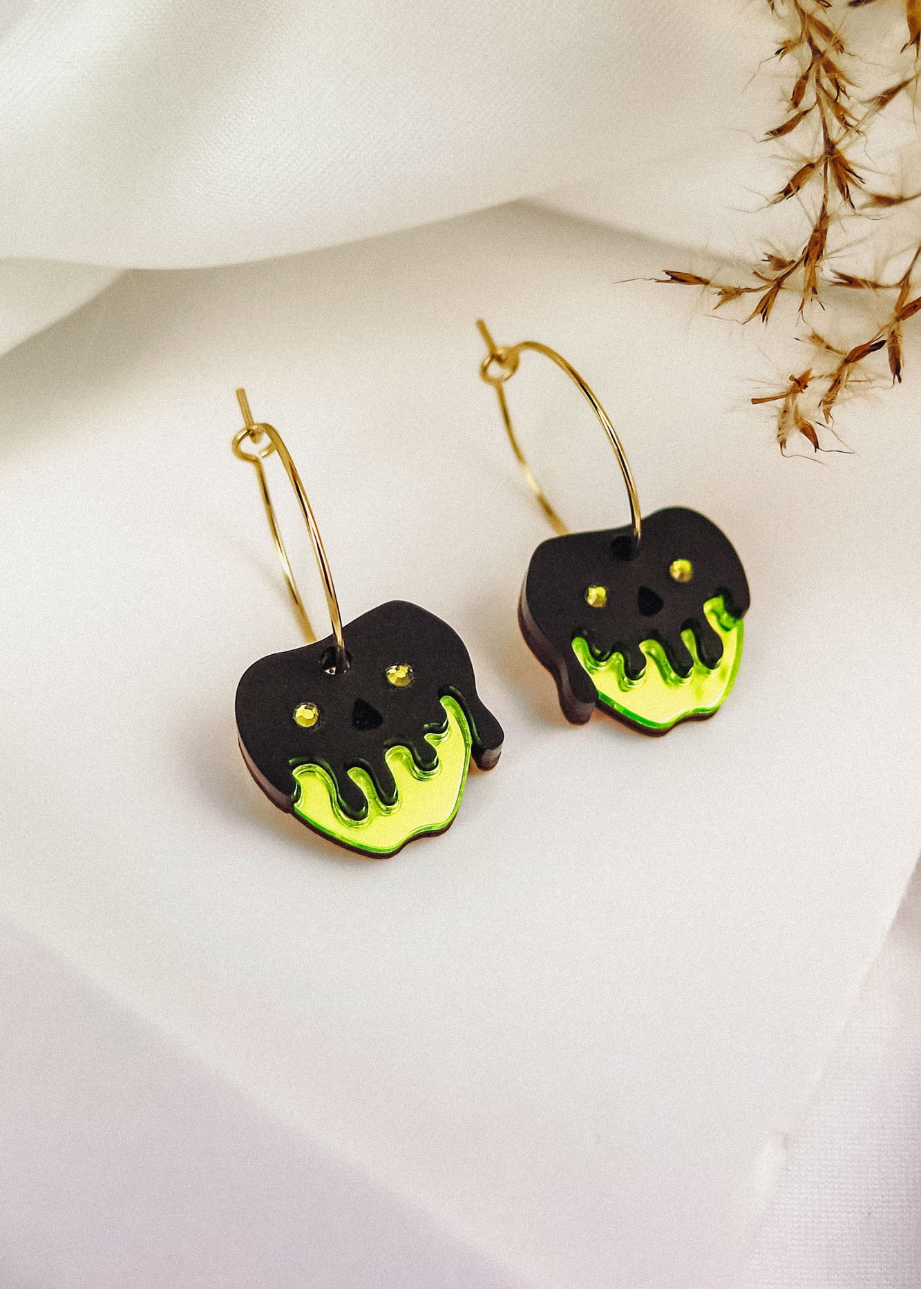 A pair of Poisoned Apple Earrings shaped like apples, with black and neon green dripping designs resembling poison, hang from gold hoops on a white surface with dried grass in the background—a limited edition Halloween design.