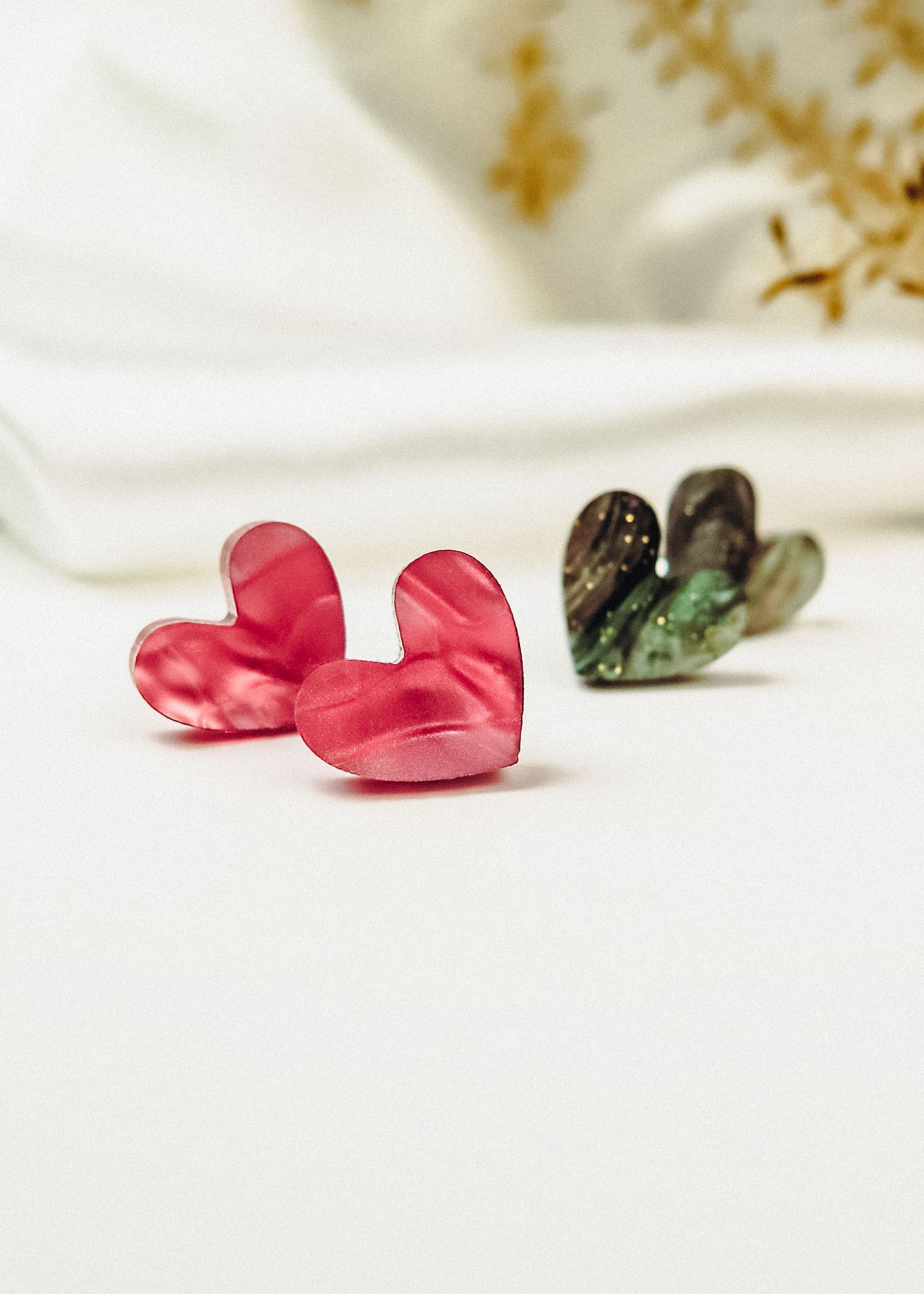 A pair of pink Tiny Heart Studs and dark green heart-shaped earrings are displayed on a white surface, radiating minimalist charm against a blurred white and yellow floral background. Hypoallergenic design ensures comfort for sensitive ears.