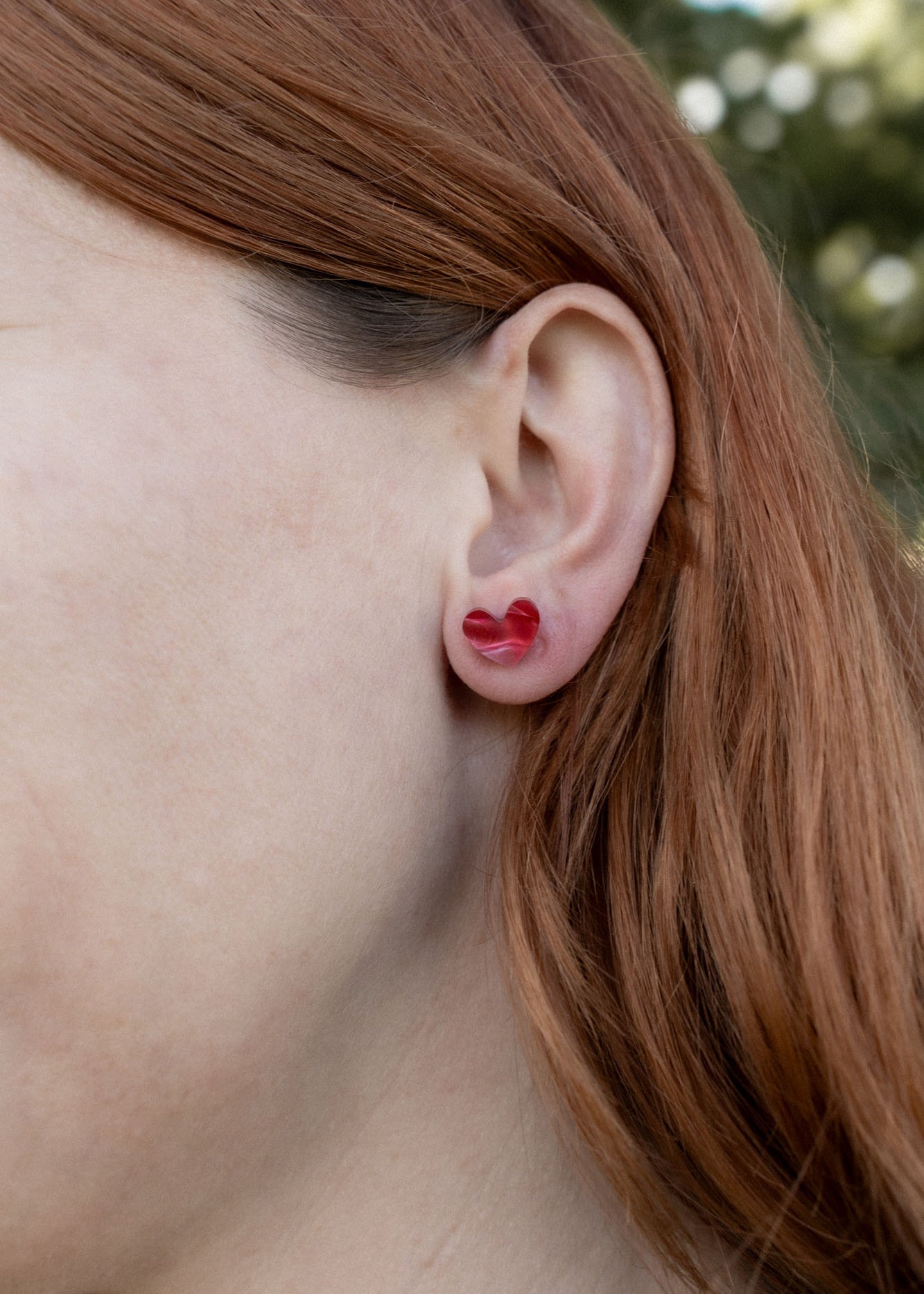 Close-up of a person with long reddish-brown hair wearing a red Tiny Heart Stud in their left ear, highlighting its minimalist charm. The background is blurred greenery.