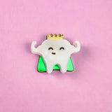 A cute Tooth King Pin enamel pin features a smiling tooth with a gold crown, arms raised in front of a green triangle on a pink background. This handcrafted accessory is perfect for adding royal flair to your collection.