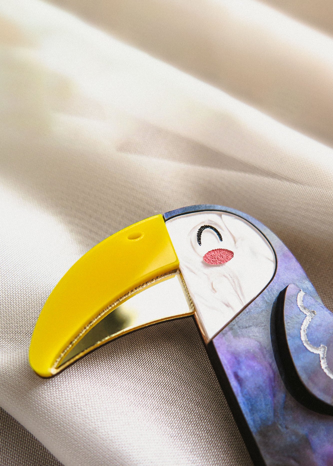 A close-up of a handcrafted Toucan Brooch with a yellow beak, white face, and vibrant painted details, resting on a soft, light-colored fabric background, radiating tropical charm.