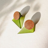 A pair of minimalist triangle earrings with round walnut wood tops and green acrylic glass bases are displayed on a light textured surface, casting soft shadows.