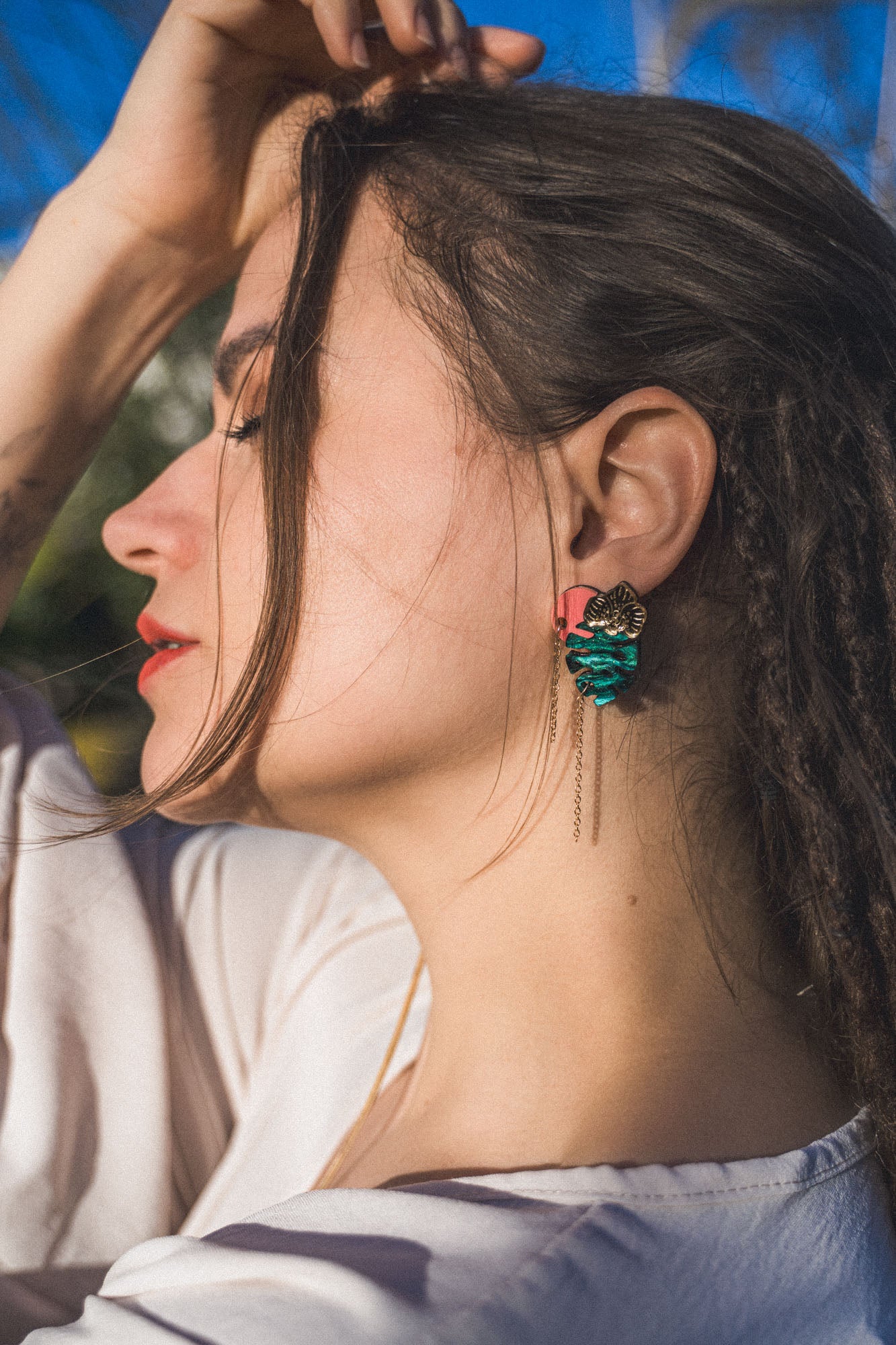 A woman with long brown hair and closed eyes tilts her head back, wearing a white top and handcrafted Tropicana Earrings featuring a tropical-inspired design with teal and gold details. Sunlight highlights her face and hair.