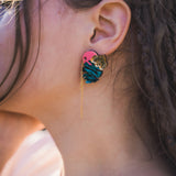 Close-up of a person’s ear wearing Tropicana Earrings—a tropical-inspired design with a gold tiger head, blue and pink details, and a delicate gold chain. The person’s hair is partially braided.