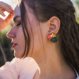 A woman with long brown hair in braids is shown in profile outdoors, wearing a pink top and handcrafted Tropicana Earrings with a tropical-inspired design shaped like leaves and a sun. Sunlight highlights her face and hair.