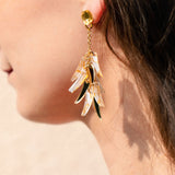Close-up of a woman’s ear wearing featherlight dangle earrings with gold, green, and translucent acrylic tufts shaped like leaves. The softly blurred background highlights the jewelry and the side of her face and hair.