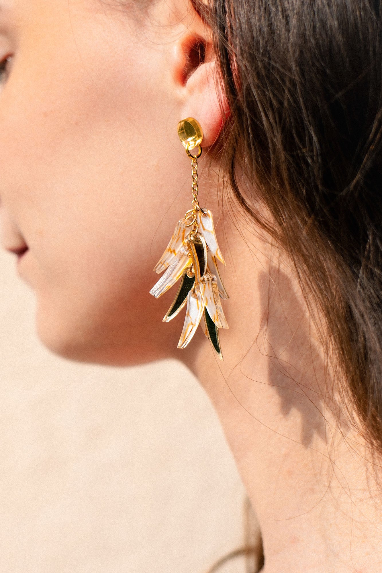 Close-up of a woman’s ear wearing featherlight dangle earrings with gold, green, and translucent acrylic tufts shaped like leaves. The softly blurred background highlights the jewelry and the side of her face and hair.