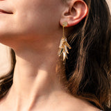 Close-up of a woman with long brown hair, eyes closed, and a slight smile, wearing featherlight gold dangle earrings shaped like cascading leaves. She is dressed in a white top as sunlight highlights her skin.