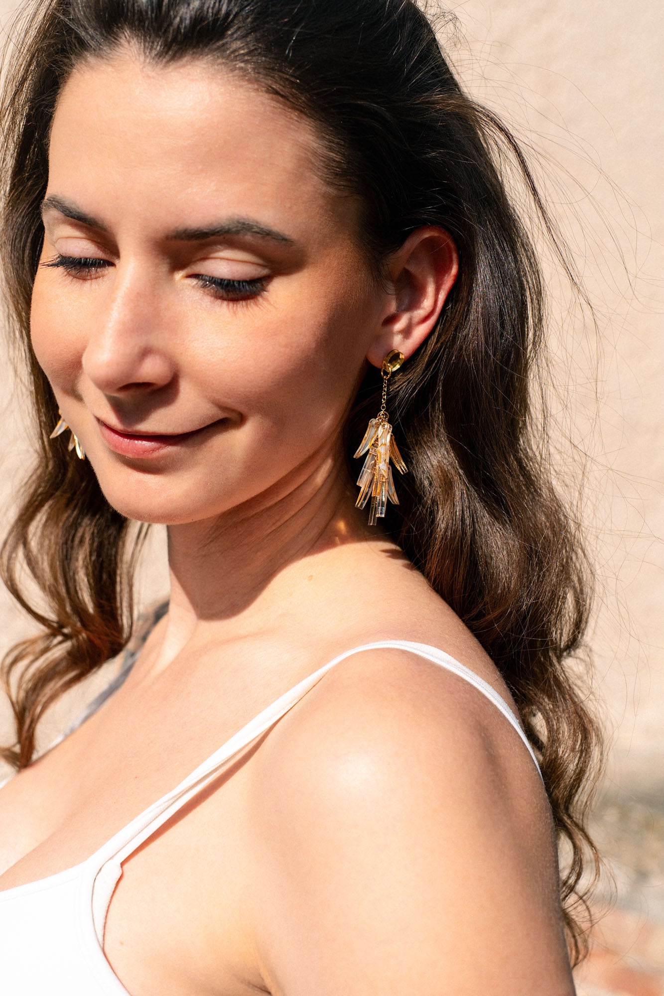 A woman with long brown hair and a white sleeveless top smiles gently with her eyes closed. She wears featherlight gold and crystal dangle earrings, standing in sunlight against a light-colored background.