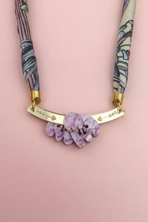 A handmade necklace featuring a pink and purple patterned fabric strap, a gold curved bar with small heart cutouts, and a cluster of marbled lavender decorative stones, displayed on a light pink background.