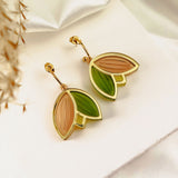 A pair of handcrafted gold outline earrings with leaf-shaped pendants in light pink and green enamel, displayed on a white card over satin fabric with dried grass in the corner. These lightweight earrings add a touch of elegance to any look.