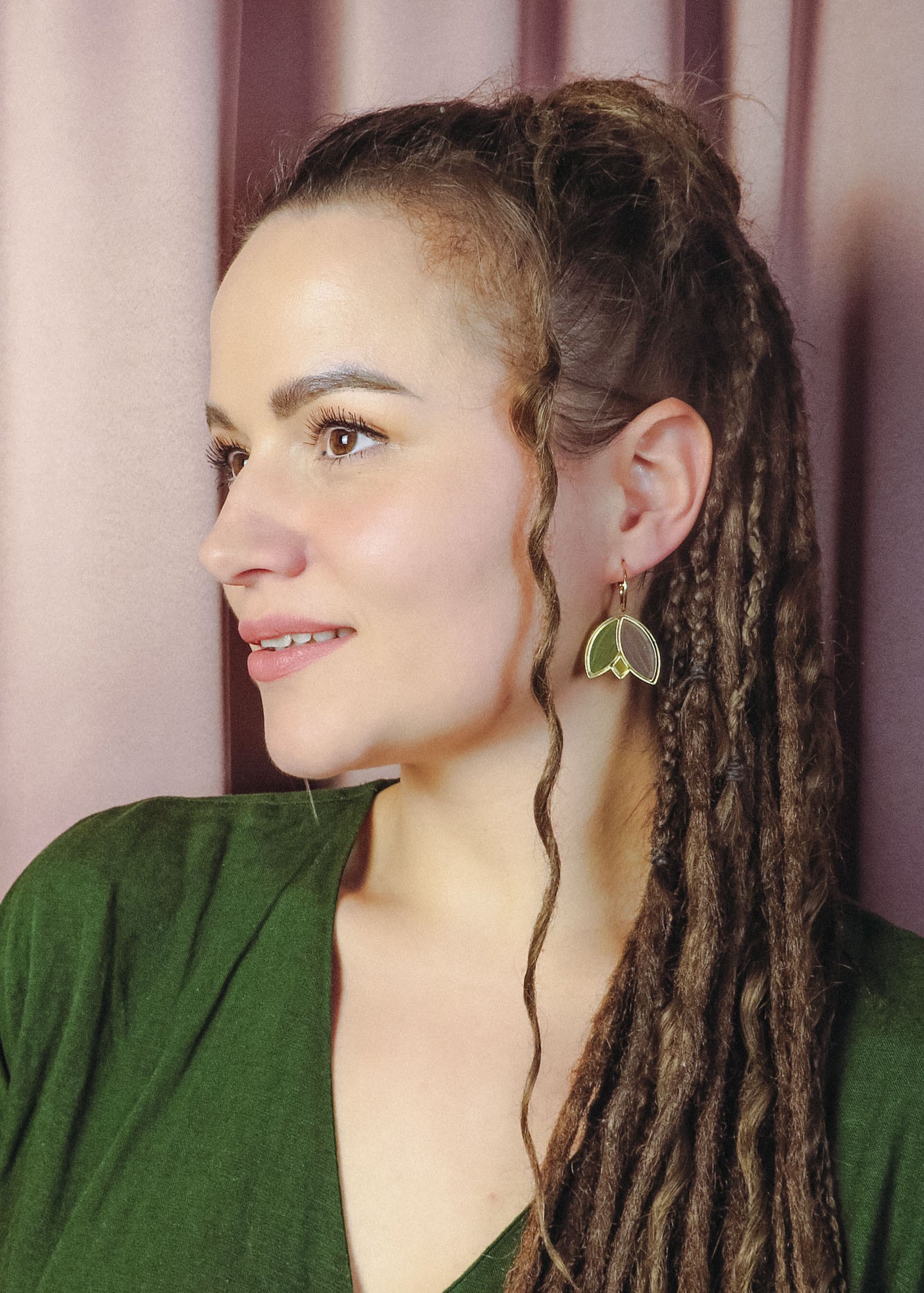A woman with long, brown dreadlocks and a loose curl framing her face wears handcrafted leaf-shaped earrings and a green top, gazing to the left against a backdrop of soft pink curtains.