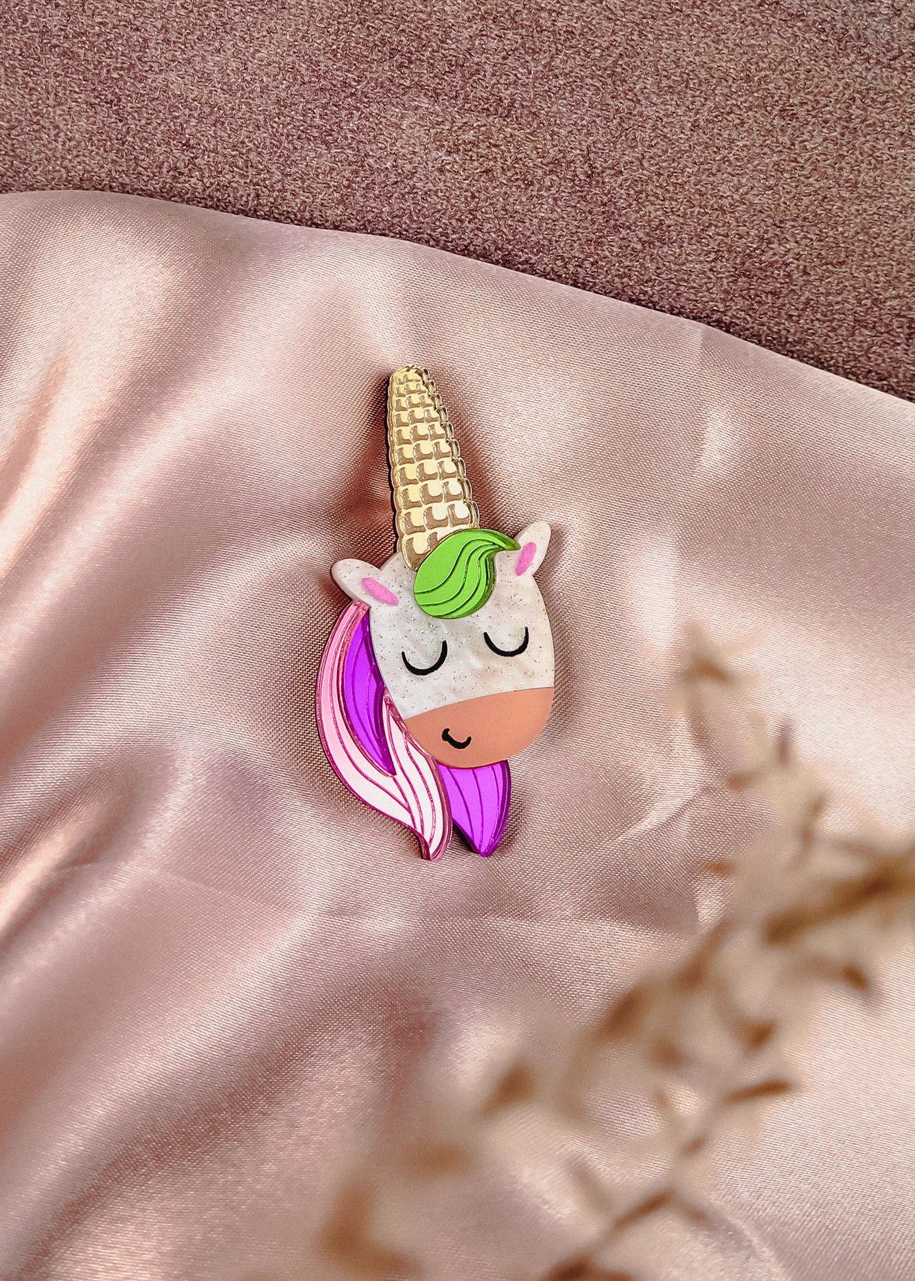 A handcrafted UniCorn Brooch with a gold horn, green and pink mane, and closed eyes rests on soft, shiny pink fabric. A blurred plant is visible in the foreground, highlighting this playful accessory’s charm.