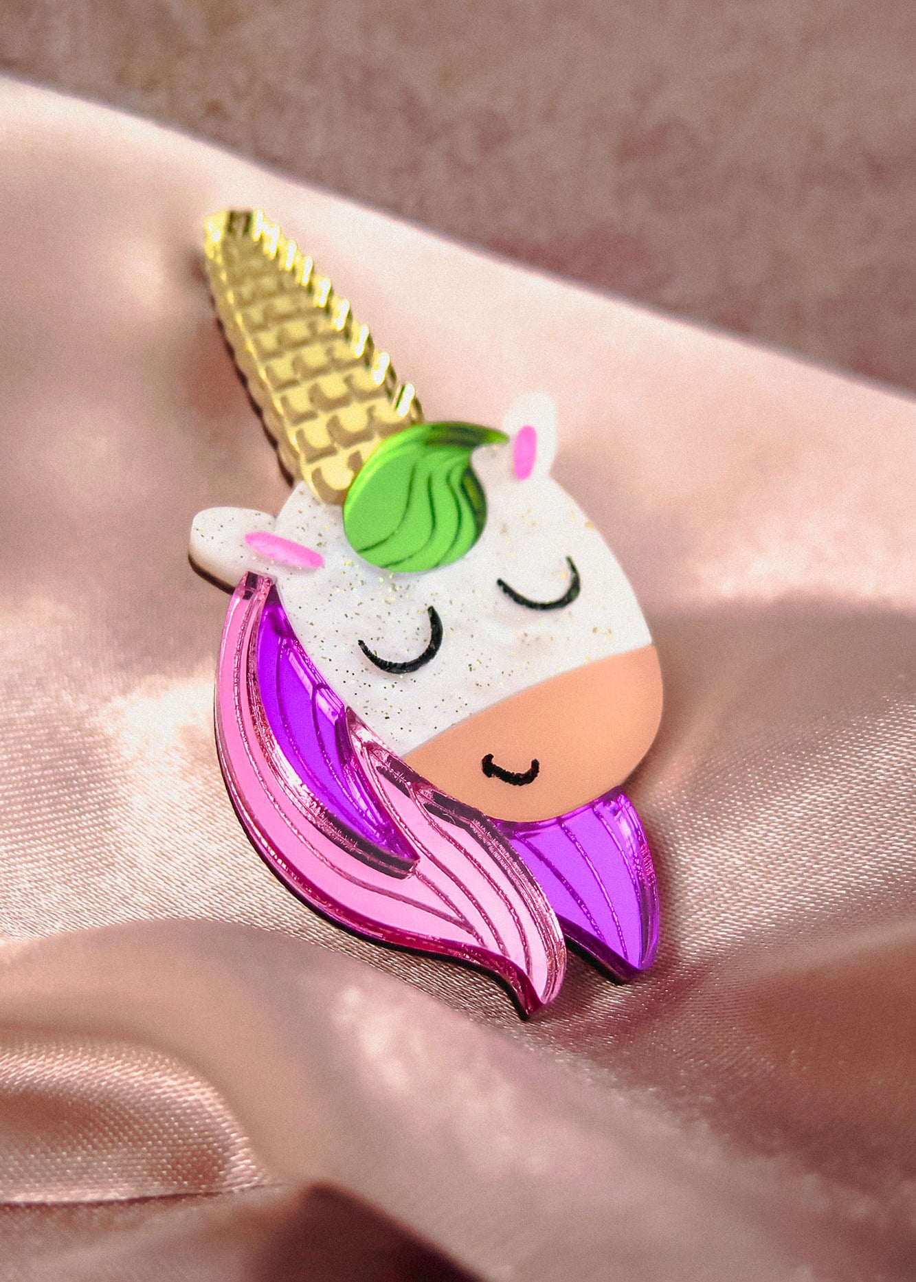 A colorful handcrafted UniCorn Brooch shaped like a unicorn’s head with a golden horn, green mane, closed eyes, and pink and purple details rests on shiny light pink satin fabric—a playful accessory for any collection.