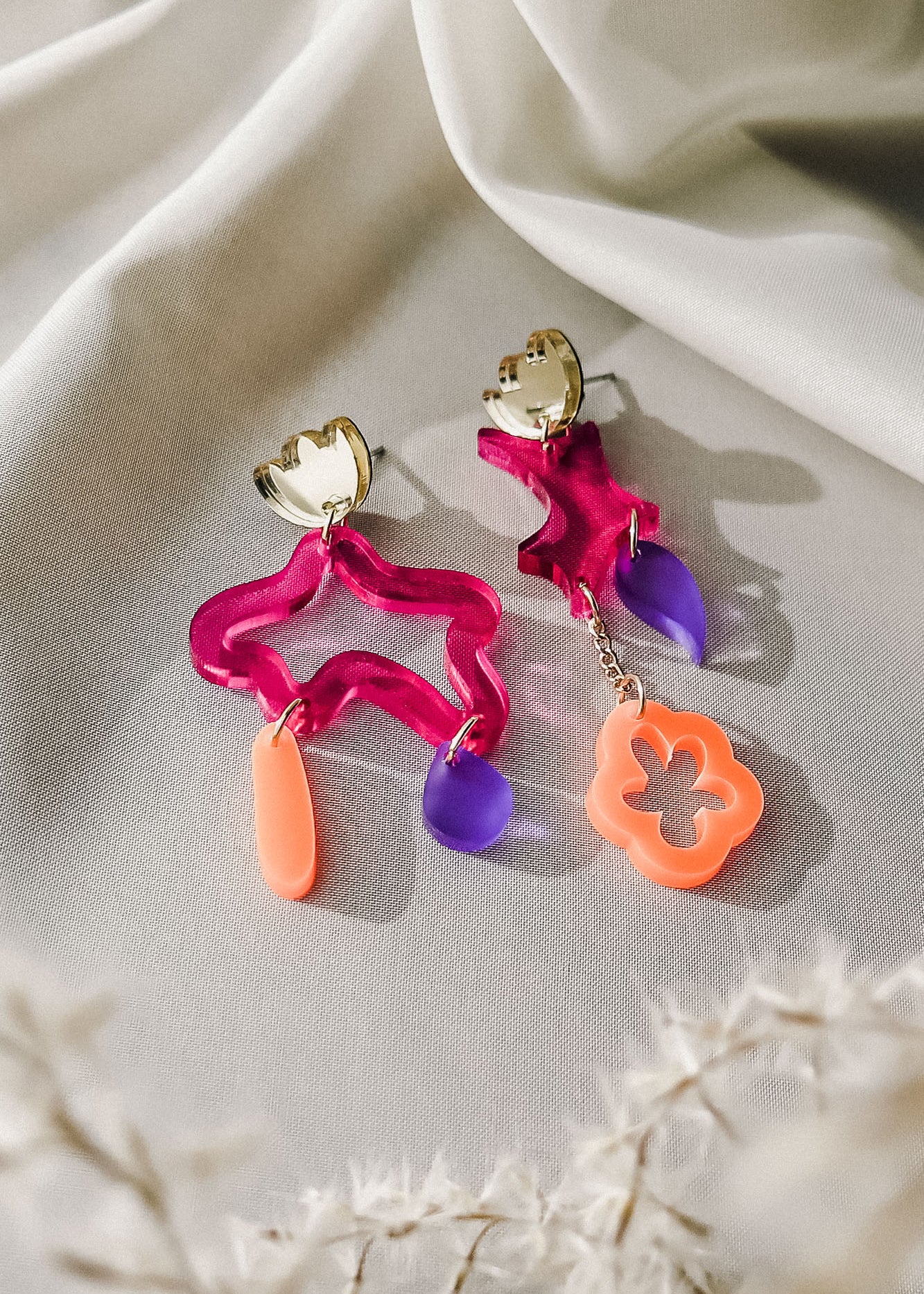Two colorful, abstract earrings with bold colors rest on soft white fabric. Each earring features gold hardware and abstract shapes in wavy pink acrylic, translucent purple, and coral orange—making this a striking pair of mismatched earrings. Dried white plants appear nearby.