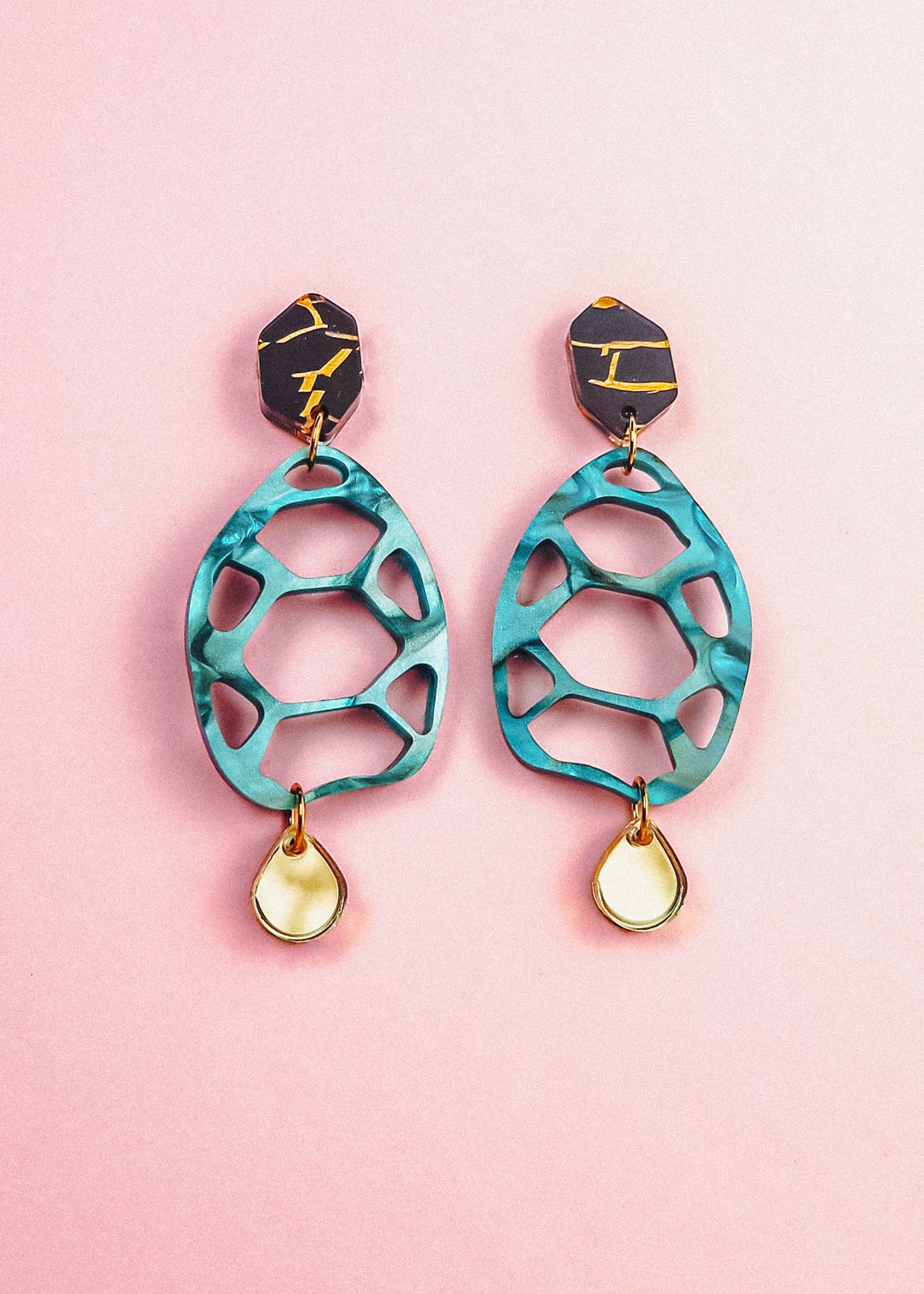 A pair of colorful, sustainable earrings with black and gold geometric tops, teal cut-out oval centers, and small teardrop-shaped gold and white dangles, displayed on a pink background—perfect for fans of handmade accessories.