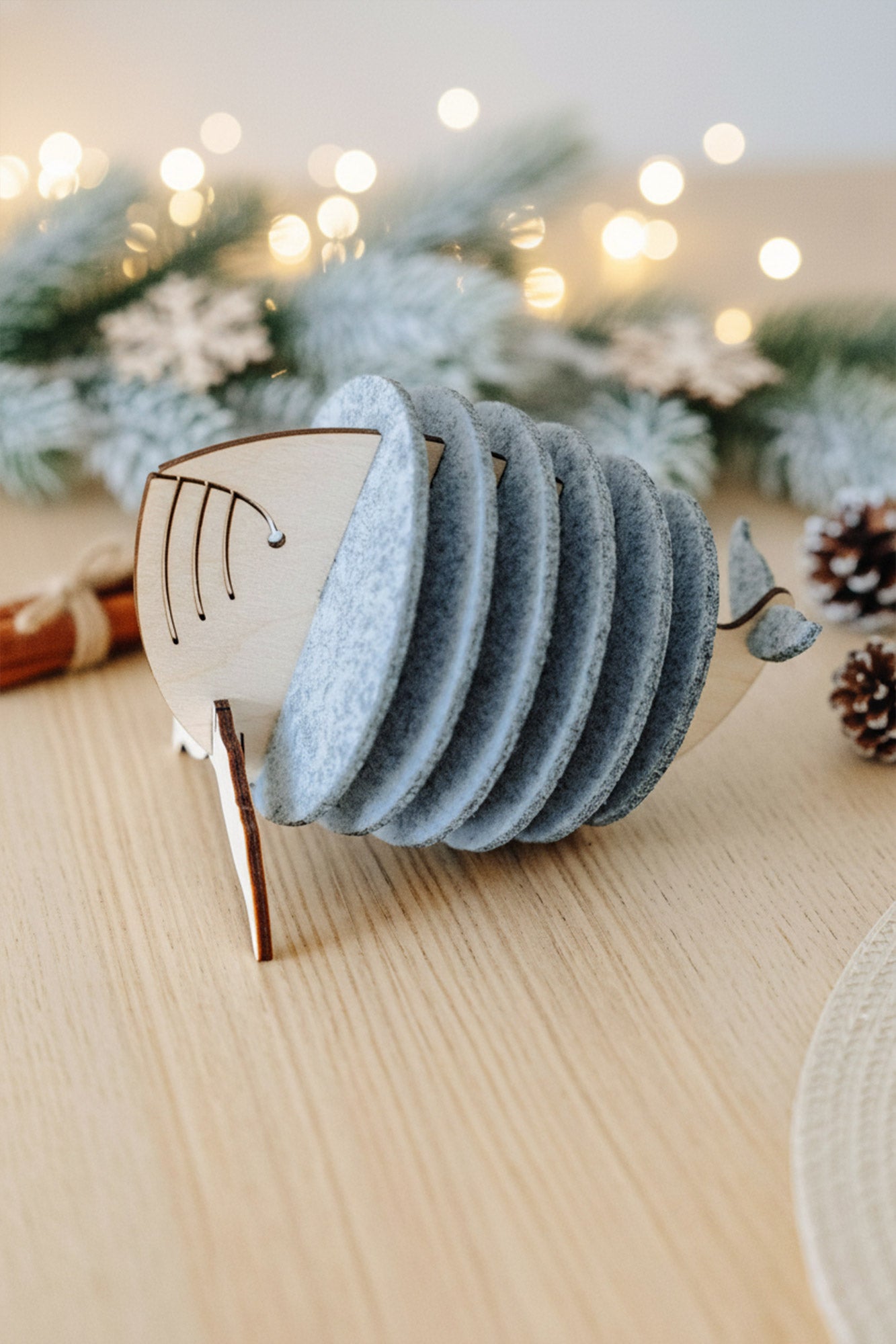 A set of cute coasters held in a wooden holder shaped like a whale, placed on a light wooden table with pine cones, fir branches, and warm fairy lights in the background—a charming touch for your home decor.