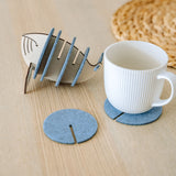 A white ribbed ceramic mug sits on a round gray felt coaster on a wooden table, with Whale Coasters and a whale-shaped holder nearby. A woven placemat adds home charm while these coasters help protect surfaces in style.