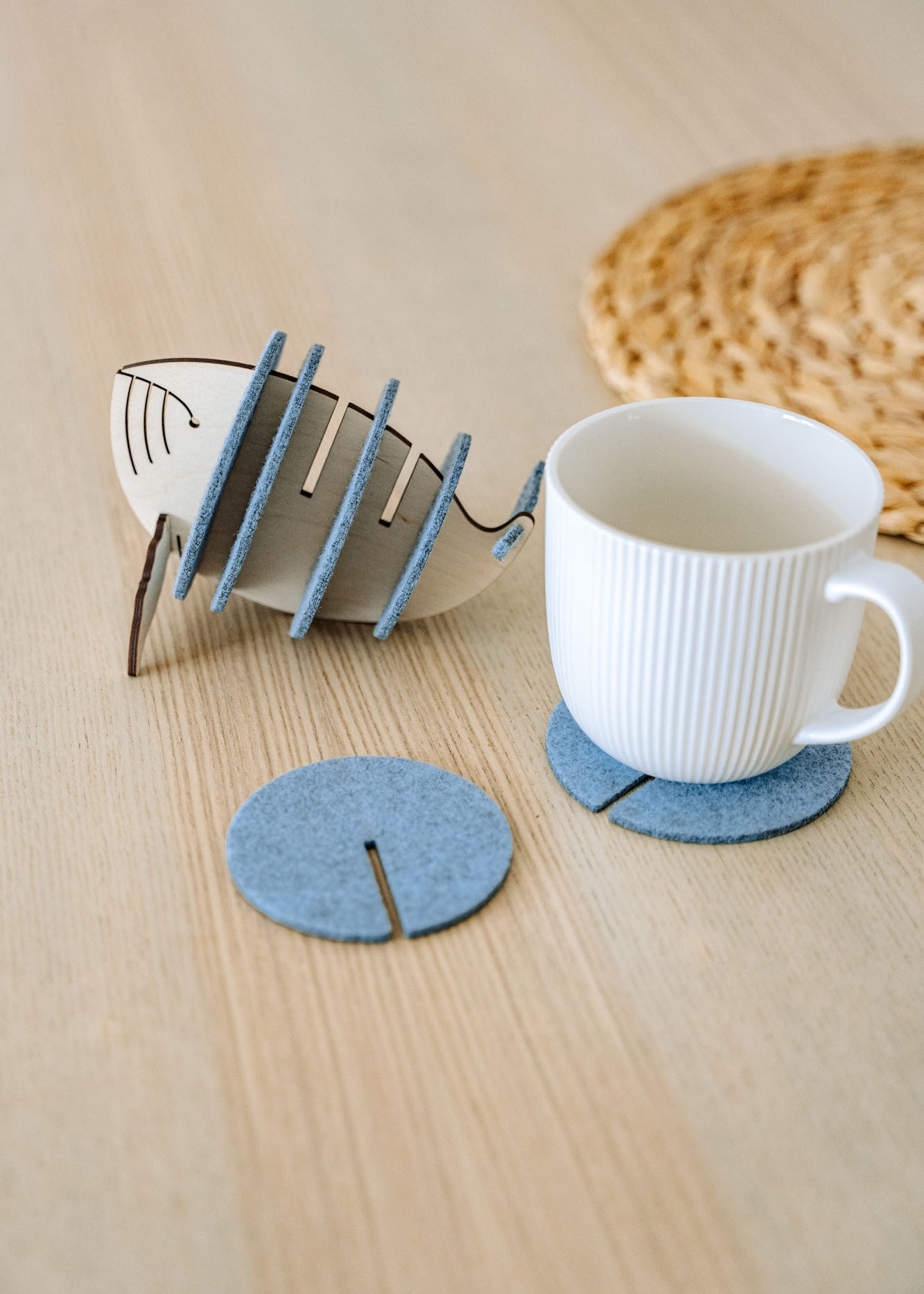 A white ribbed ceramic mug sits on a round gray felt coaster on a wooden table, with Whale Coasters and a whale-shaped holder nearby. A woven placemat adds home charm while these coasters help protect surfaces in style.
