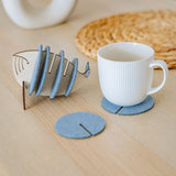 A white ribbed mug sits on a round gray felt coaster, designed to protect surfaces. Nearby, more Whale Coasters are stacked in a fish-shaped holder, with woven and ceramic decor adding home charm in the background.